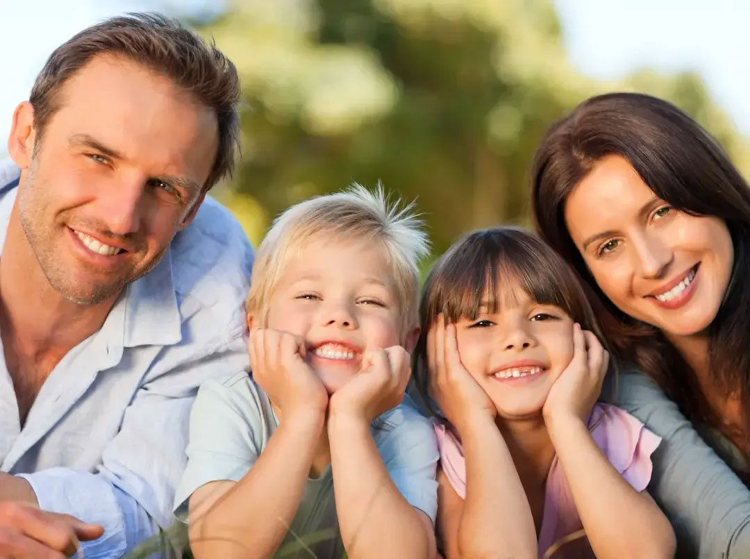 Family of four, including two adults and two children, posing for a photo with smiles, set against a blurred outdoor background.