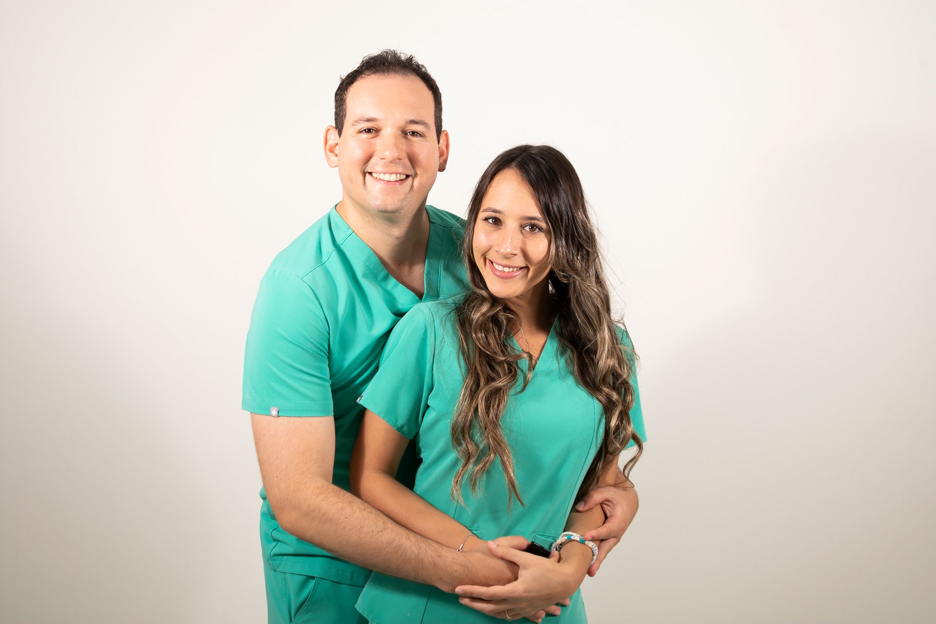 A man and a woman, both dressed in scrubs, are posing for a photograph with one arm around each other.