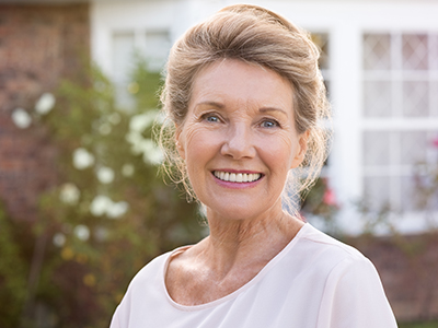 The image is a portrait of an older woman with short blonde hair, wearing makeup and a white top, smiling and looking directly at the camera. She has a fair complexion and appears to be outdoors, possibly in front of a house or garden, as suggested by the presence of greenery and a brick structure in the background.