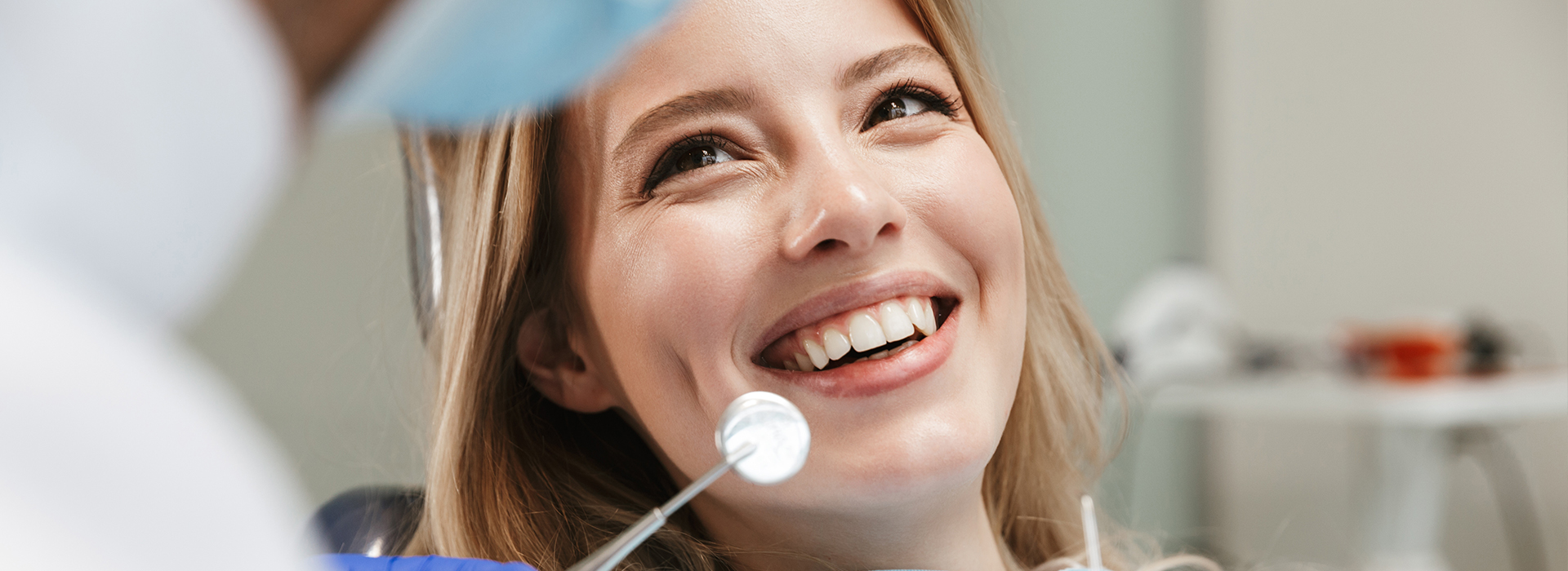 A woman smiling at a camera with a dental professional in the background, both in a dental office setting.