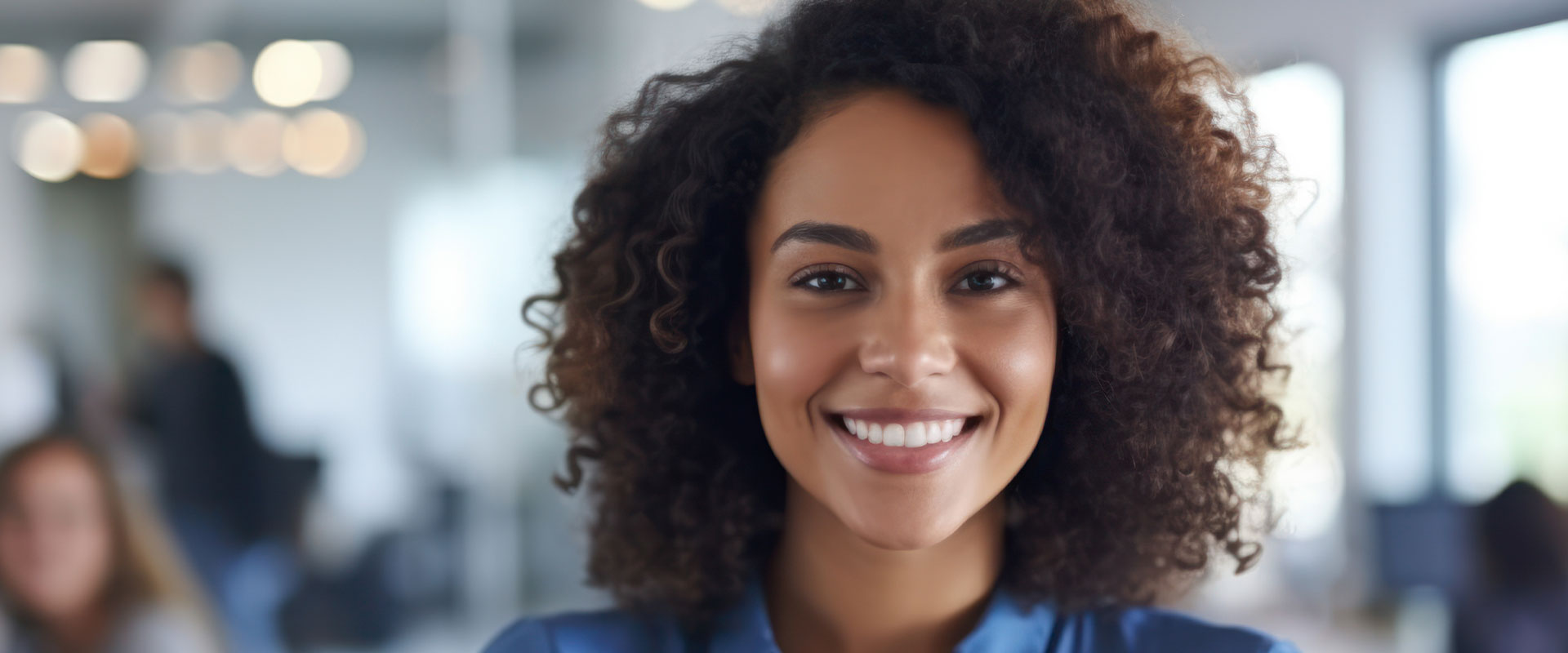 The image features a smiling woman with curly hair, wearing a dark top and seated in an office environment.