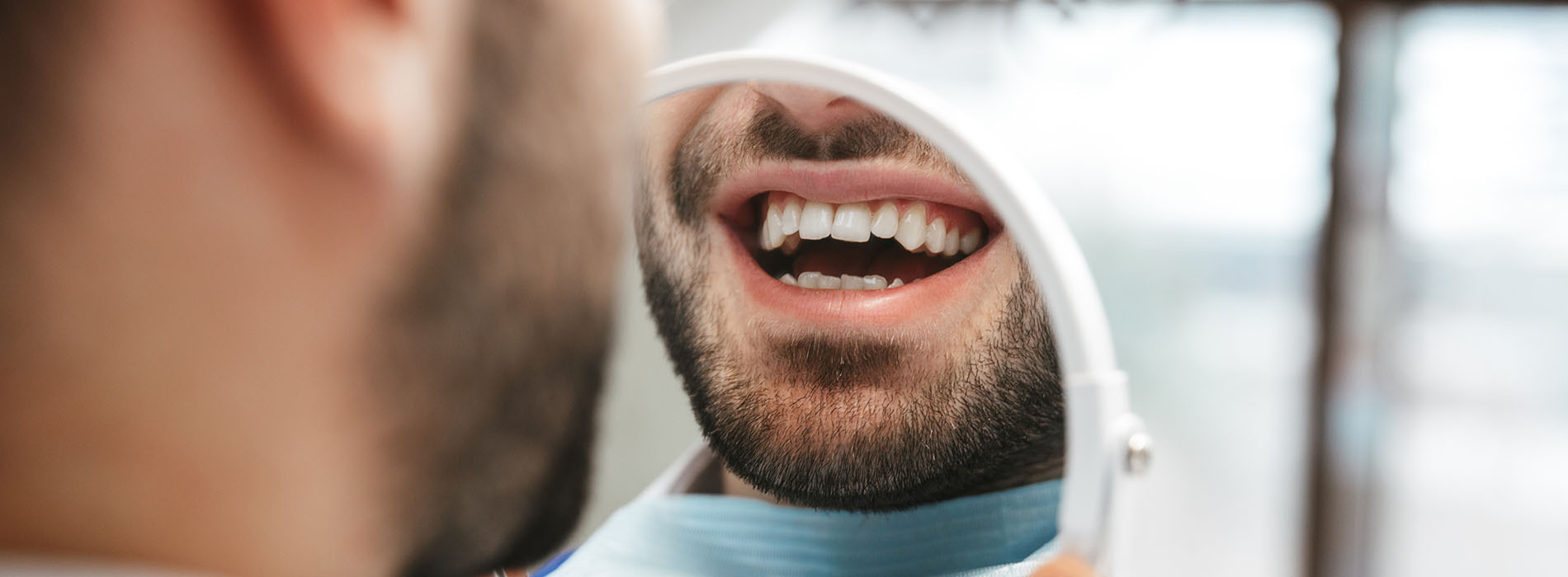 The image shows a man with a beard sitting in a dentist s chair, smiling at the camera while having his teeth cleaned.