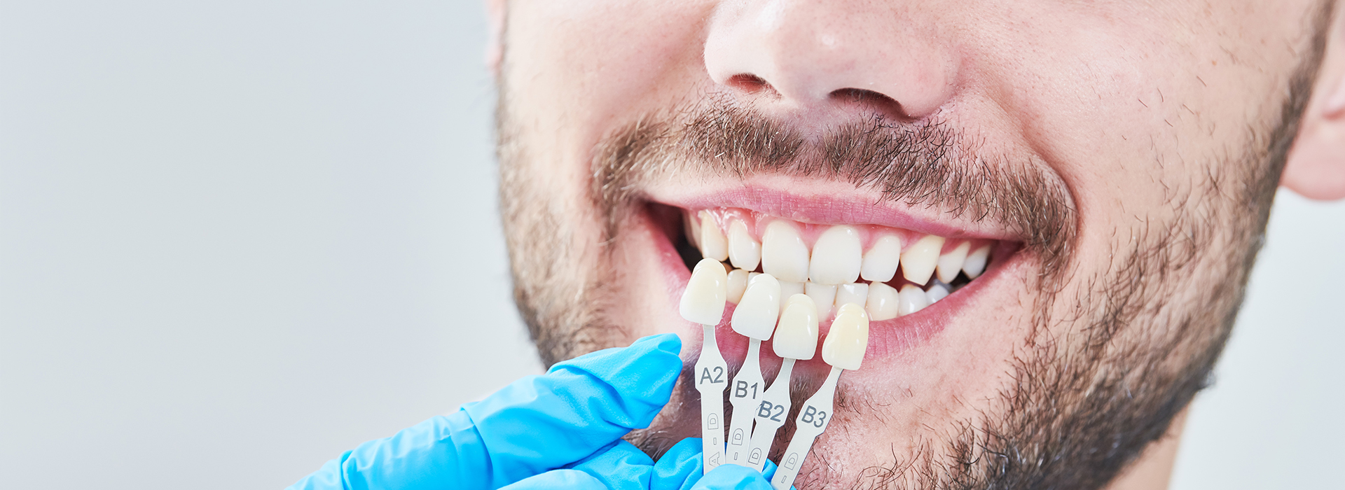 The image shows a man with a toothy smile, wearing a blue surgical mask and holding dental instruments.