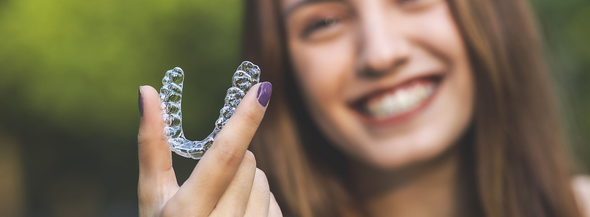 An image of a person smiling, holding up a clear plastic retainer with one hand.